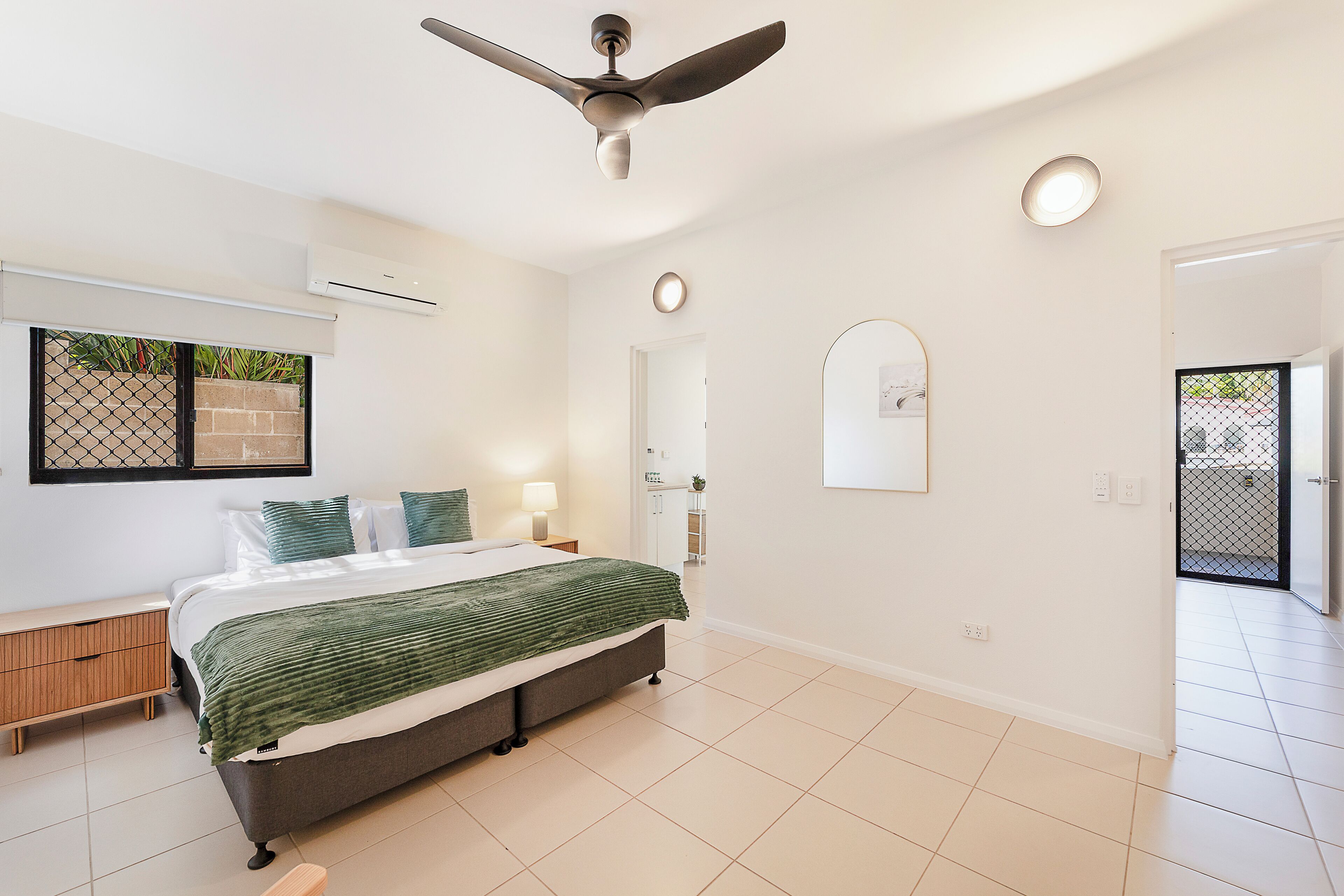 A bedroom with a bed, bedside tables, a ceiling fan, an air conditioning unit, a window with a view, a door, and a tiled floor.