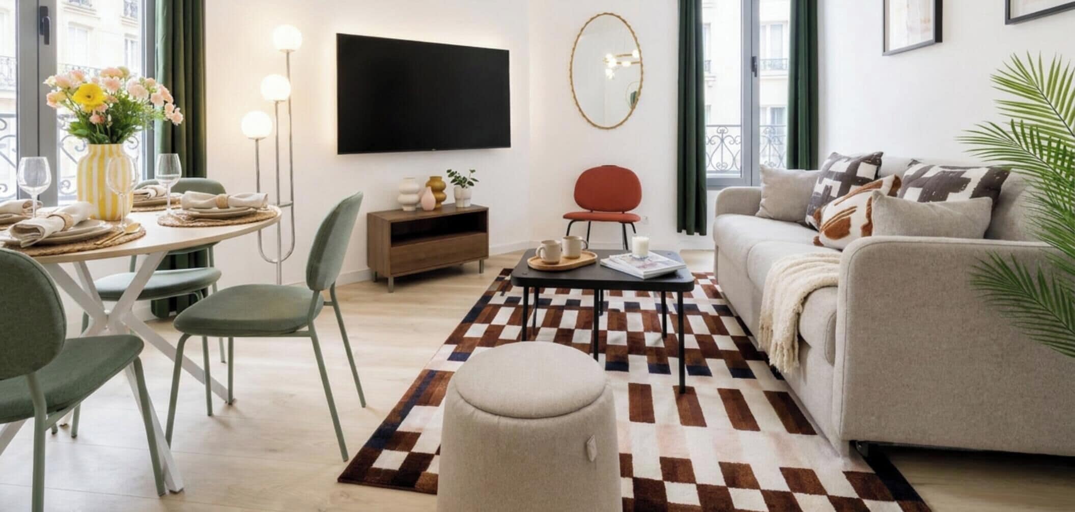 A modern living room with a sofa, a dining table with chairs, a coffee table, a TV mounted on the wall, and a patterned rug.