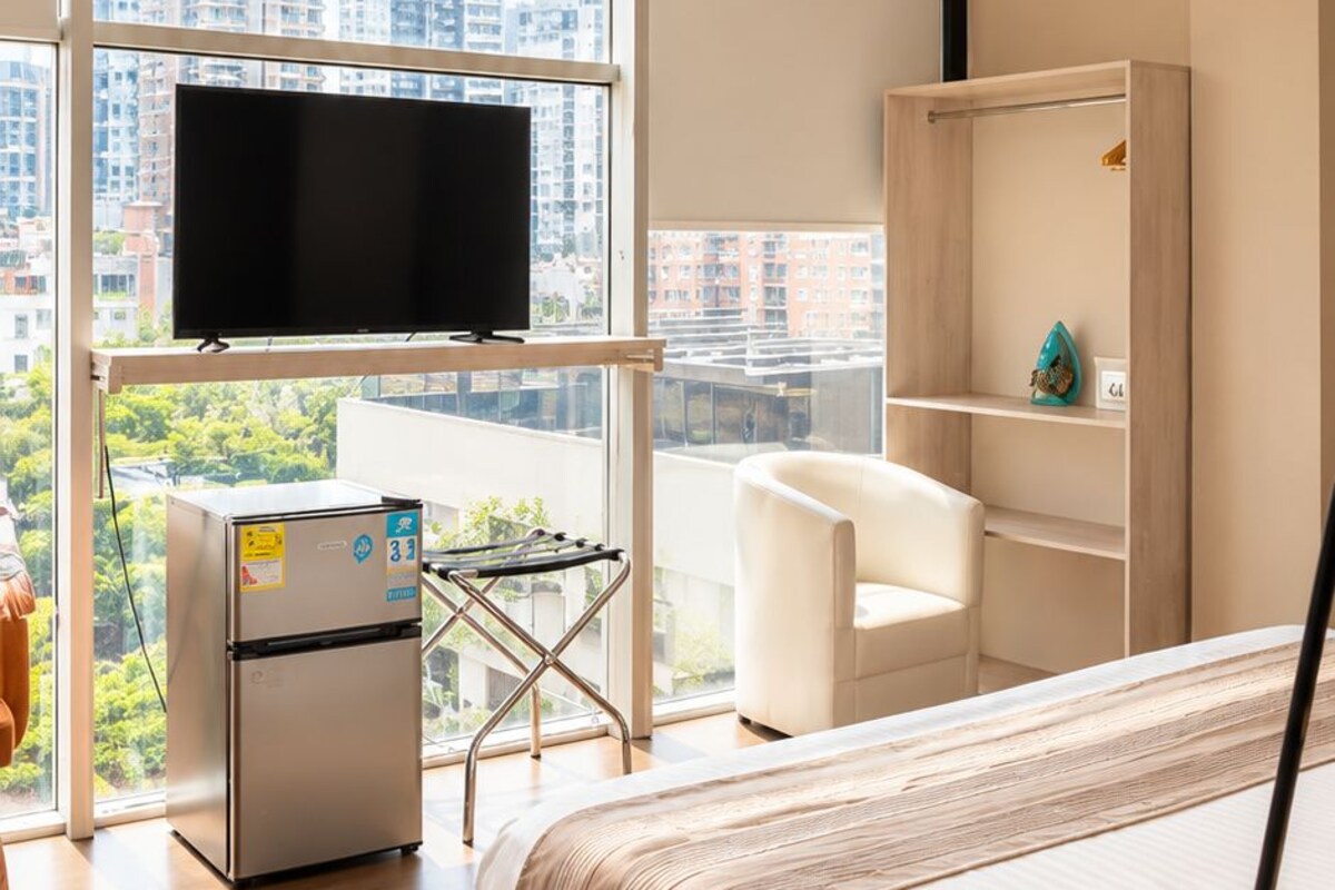 Habitación de hotel moderna con una cama grande, un televisor de pantalla plana, un refrigerador pequeño, un escritorio con una silla y vistas a la ciudad a través de amplias ventanas.