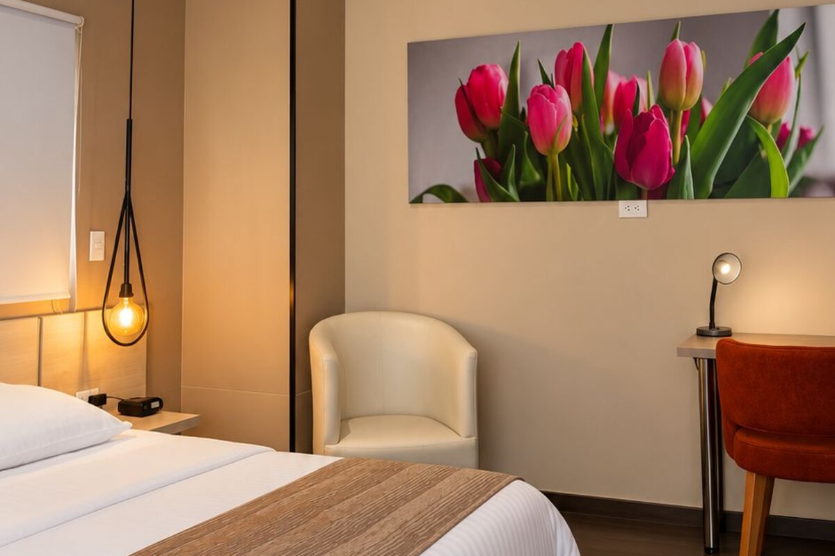 Habitación de hotel con cama, una silla, una mesita, un cuadro colgado en la pared y una lámpara colgante.