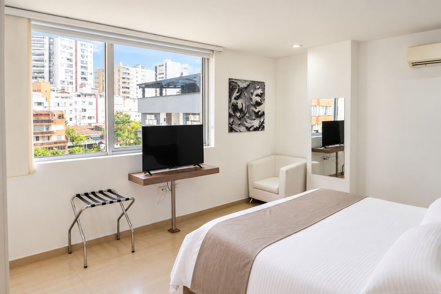A hotel room with a bed, a TV on a stand, a chair, a window with a city view, and a painting on the wall.