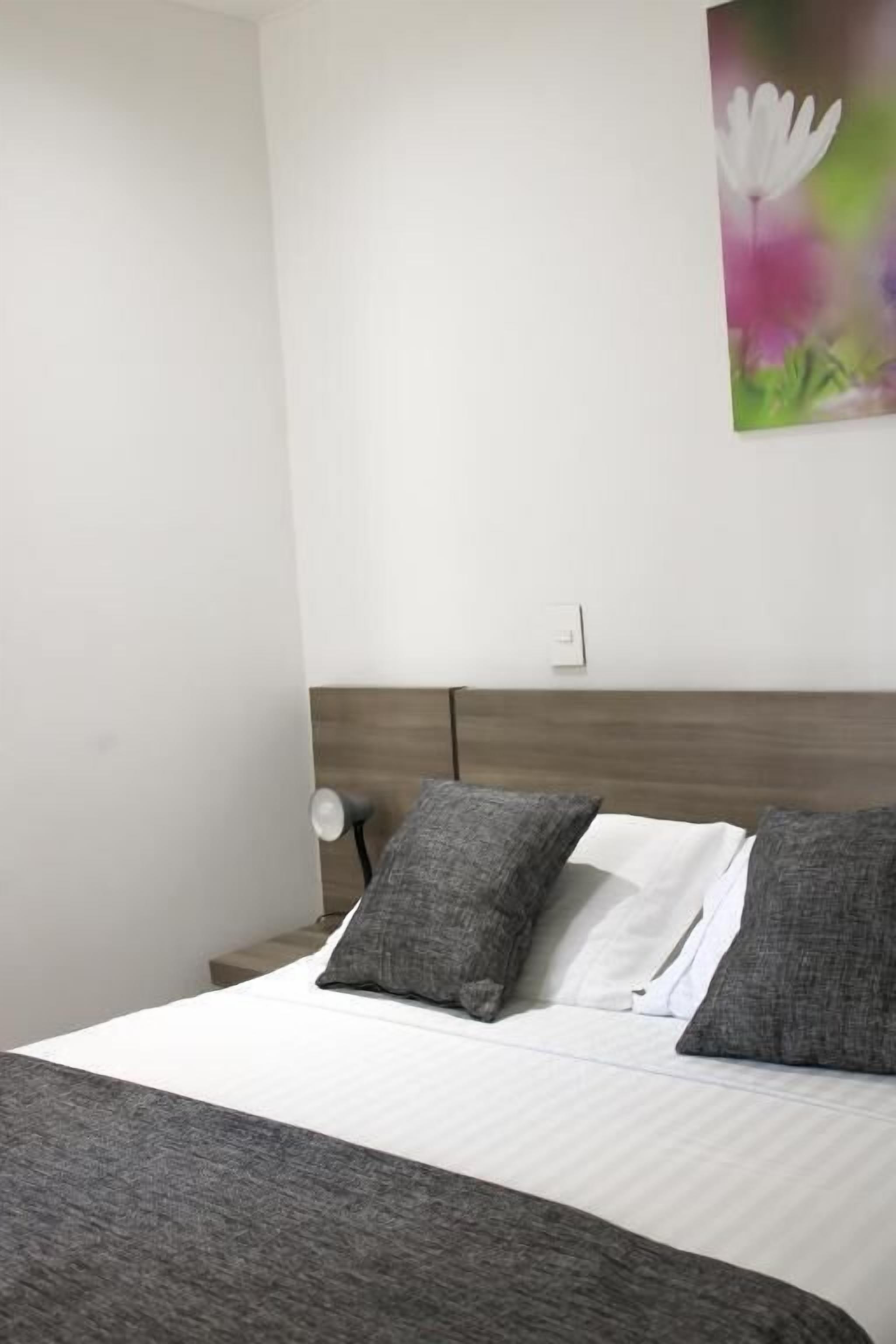 A bed with white and gray bedding, a wooden headboard, and a wall-mounted light fixture.