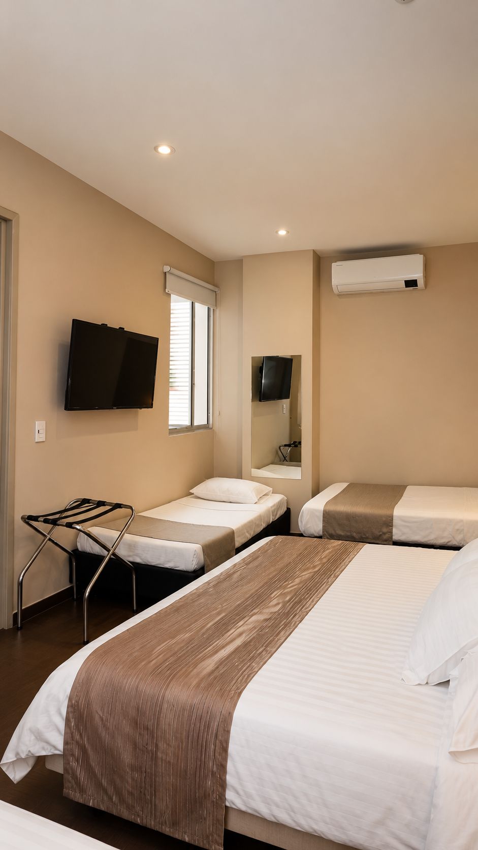 A hotel room with two beds, a TV, an air conditioning unit, and a window with blinds.