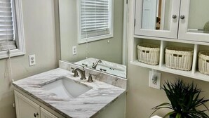 A bathroom with a marble sink, a mirror, a window with blinds, and wicker baskets on shelves. - The Escape on Blade Bay (Hardin County)