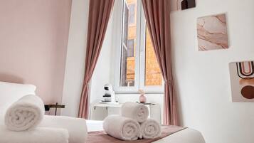 Habitación de hotel con cama, toallas enrolladas, ventana con cortinas, una mesita con un florero y decoración en la pared.