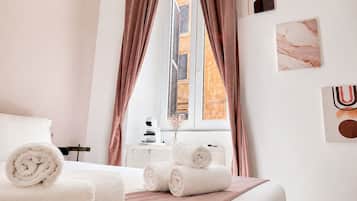Habitación de hotel con cama, toallas enrolladas, mesita de noche, ventana con cortinas y decoración en la pared.