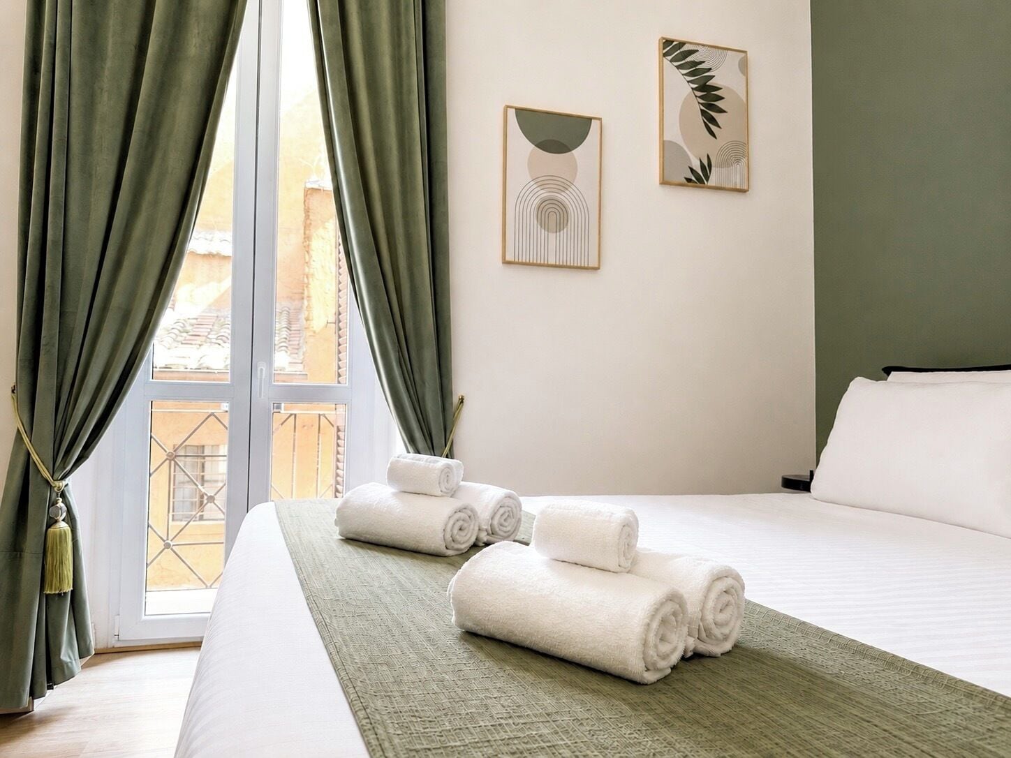 Una habitación de hotel con cama, cortinas verdes, una ventana con vistas y dos cuadros en la pared.