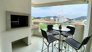 Un balcón con una parrilla, una mesa redonda con sillas y vistas a edificios y colinas.