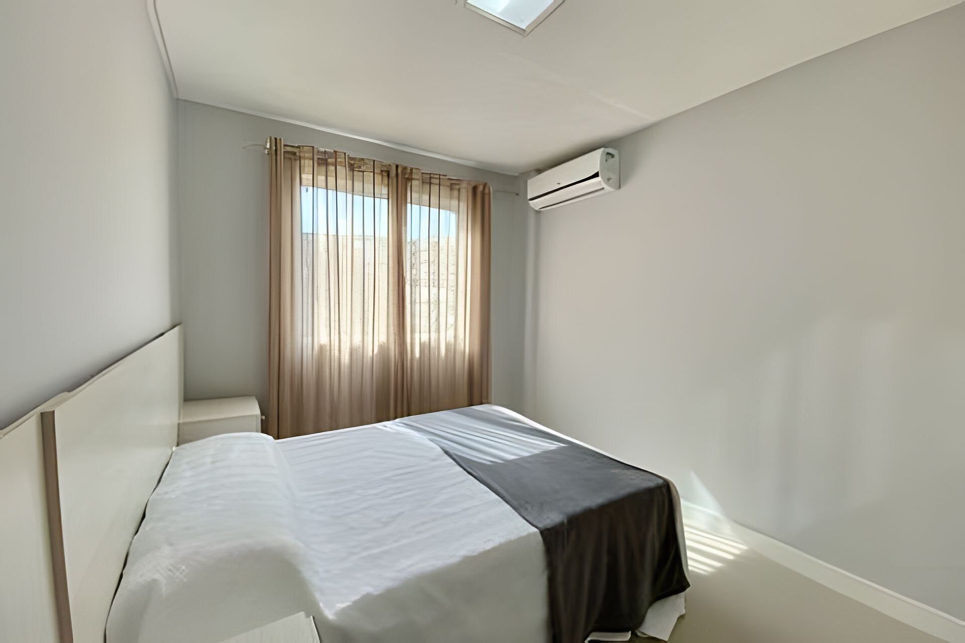A single bed with a white and black bedspread, a window with sheer curtains, and an air conditioning unit mounted on the wall.