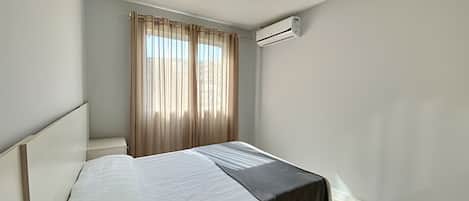 A single bed with a white and black bedspread, a window with sheer curtains, and an air conditioning unit mounted on the wall.