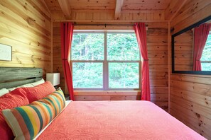 A wooden cabin bedroom with a bed, a window with curtains, and a view of the forest. - 5 Minutes to Ash Cave! Sleeps 10 w/3 Private Bedrooms + Hot Tub + WiFi + Games (South Bloomingville)