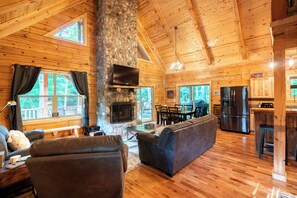 A wooden cabin interior with a stone fireplace, a dining area, and a kitchen. - 5 Minutes to Ash Cave! Sleeps 10 w/3 Private Bedrooms + Hot Tub + WiFi + Games (South Bloomingville)