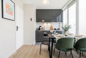 Une cuisine moderne avec des meubles de couleur sombre, une table à manger pour quatre personnes et une grande fenêtre laissant entrer la lumière naturelle.