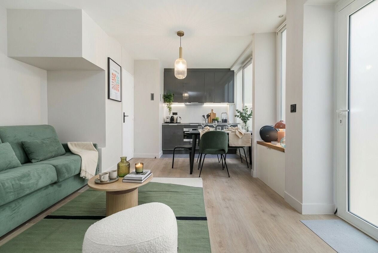 Ein modernes Wohnzimmer mit einem grünen Sofa, einem Couchtisch und einem weißen Sessel. Im Hintergrund ist eine Küche mit Essbereich zu sehen.