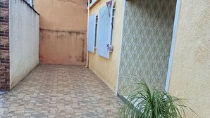 A paved courtyard with a potted plant, a tiled wall, and a window with a patterned curtain.