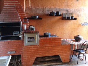 A brick oven with a metal door, a table with a patterned tablecloth, and shelves with pots and a sign. - Fully furnished house, everything included. (Betim)