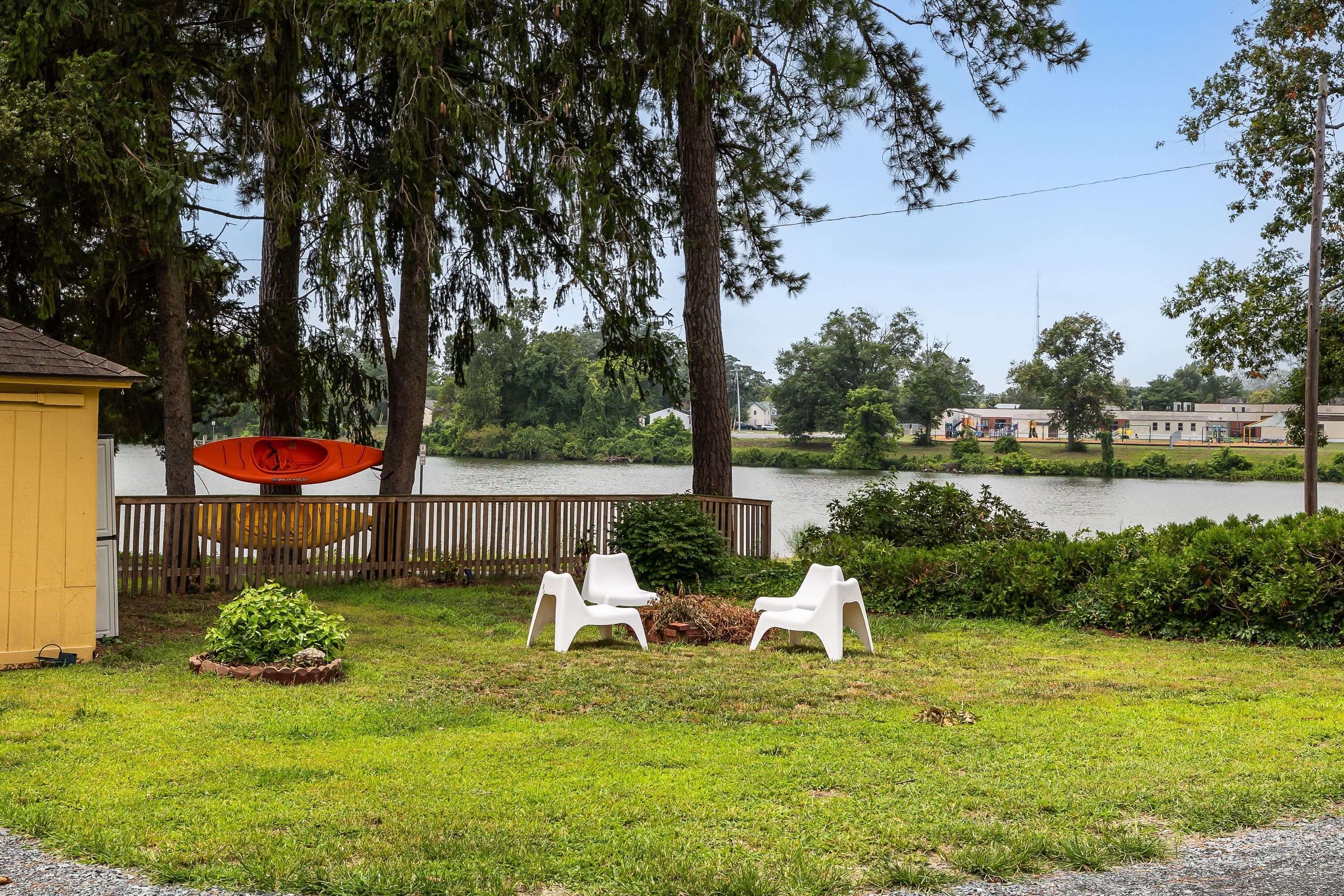A lakeside property with a fenced yard, two white plastic chairs, a red kayak, and a yellow building.