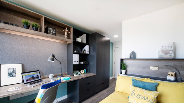 A modern bedroom with a desk, a bed with a headboard, a chair, and a shelf with decorative items. - Student Only Anglia House (Cambridge)