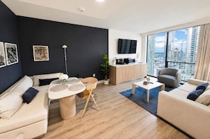 A modern living room with a sofa, a dining table with chairs, a coffee table, a TV mounted on the wall, and a large window with a city view. - 1BR with Bay Views, Pool & 24h Gym in Downtown Miami G1918 (Miami Beach)
