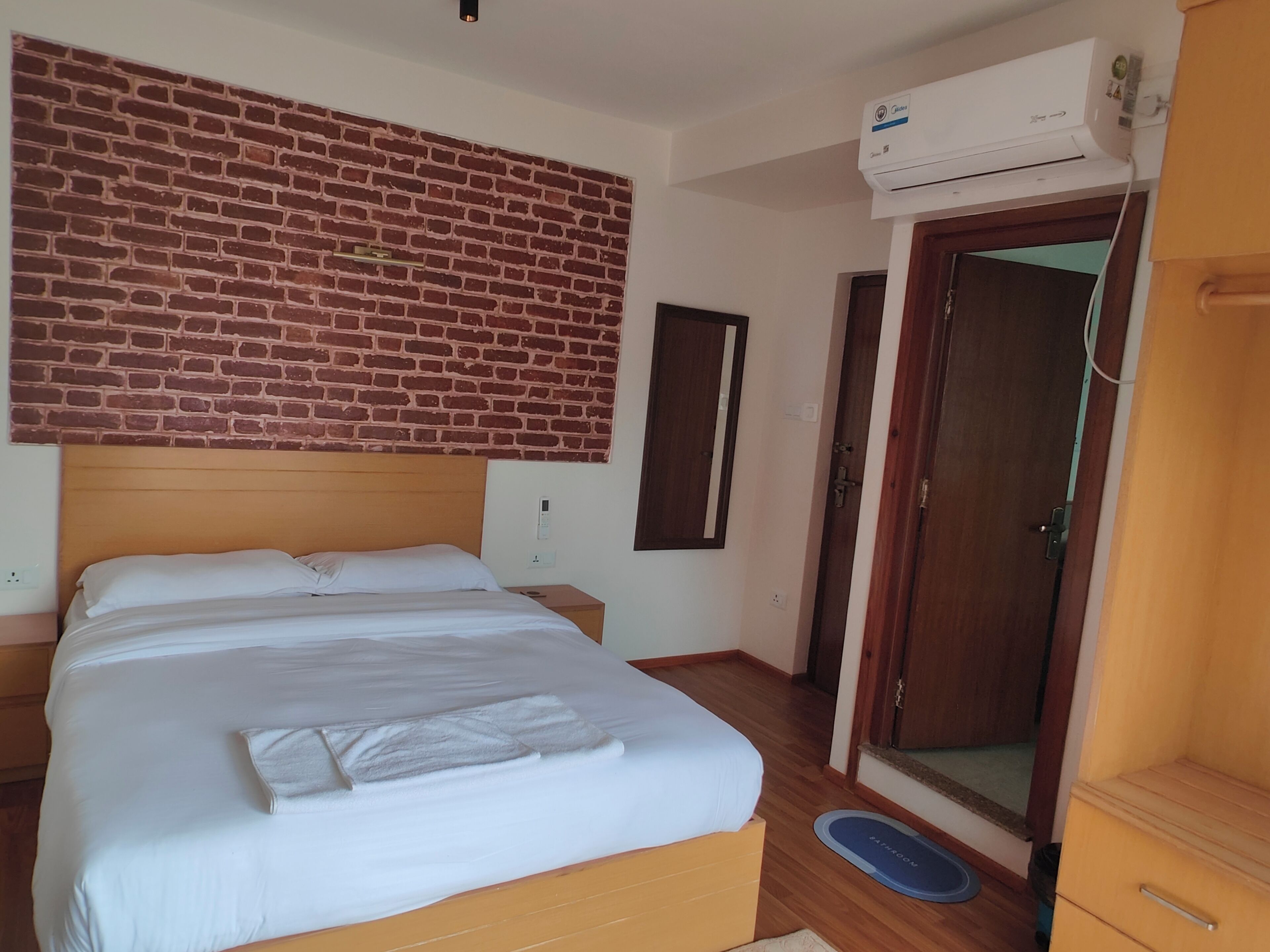 A hotel room with a brick-patterned wall, a bed with white linens, a wooden headboard, a wooden wardrobe, a door with a mirror, and an air conditioning unit.