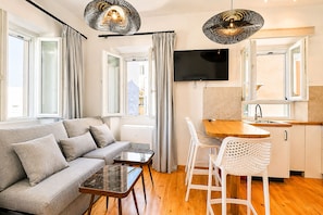 A modern living space with a sofa, a coffee table, a TV, and a kitchen area with a sink and stools. - Casa Dei Padri #2 - Historic Center (Corfu)