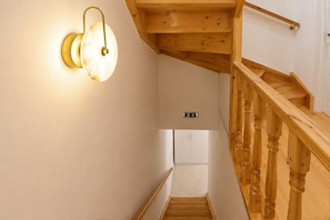 A wooden staircase with a handrail, a light fixture on the wall, and a sign indicating the direction to the exit. - Casa Dei Padri #2 - Historic Center (Corfu)