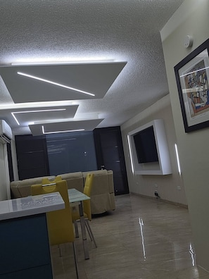 A modern interior with a dining area, a flat-screen TV mounted on the wall, and a ceiling with linear light fixtures. - Fajardo Coastal Retreat: 2BR w/ 2 Pools (Fajardo)