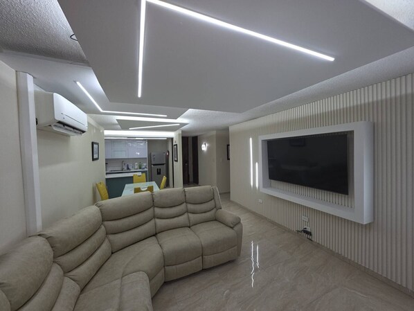 A modern living room with a sectional sofa, a flat-screen TV mounted on the wall, and a kitchen area in the background. - Fajardo Coastal Retreat: 2BR w/ 2 Pools (Fajardo)