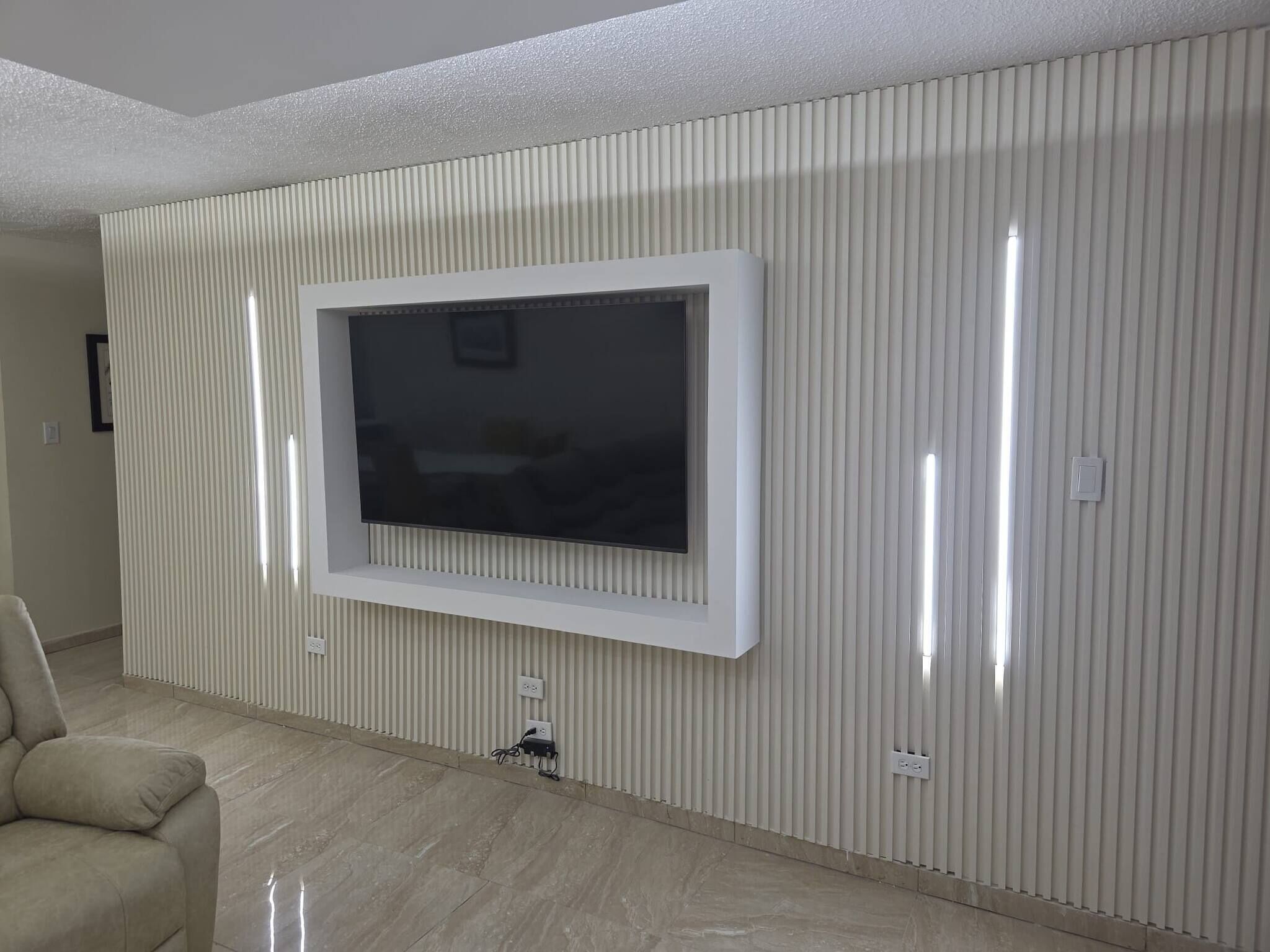 A living room with a flat-screen TV mounted on a wall with vertical paneling, a beige sofa, and a tiled floor.