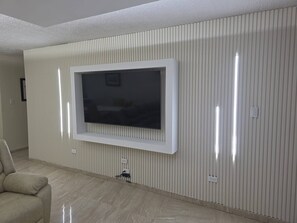 A living room with a flat-screen TV mounted on a wall with vertical paneling, a beige sofa, and a tiled floor. - Fajardo Coastal Retreat: 2BR w/ 2 Pools (Fajardo)