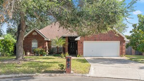 A residential house with a brick facade, a garage, and a front yard with a tree and potted plants.