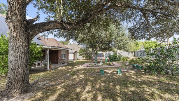 A residential backyard with a large tree, a patio with outdoor furniture, and a small garden area with a birdbath.