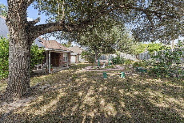 A residential backyard with a large tree, a patio with outdoor furniture, and a small garden area with a birdbath.