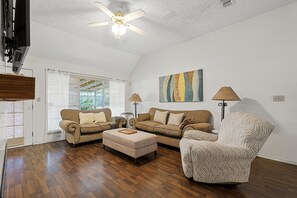 A living room with a ceiling fan, two sofas, a patterned armchair, and a painting on the wall.