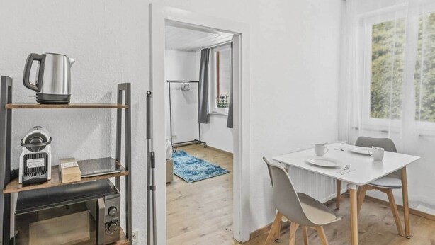 Et moderne køkken med elkedel, brødrister og ovn. Et spiseområde med bord og stole kan ses igennem en åben dør.