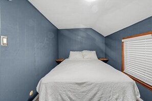 A bedroom with a single bed, a nightstand, a window with blinds, and a wall switch.