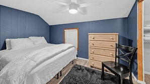 Une chambre à coucher comprenant un lit, une commode en bois, une chaise, un ventilateur de plafond et une fenêtre avec des stores.