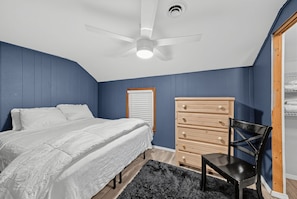 A bedroom with a bed, a wooden dresser, a chair, a ceiling fan, and a window with blinds.