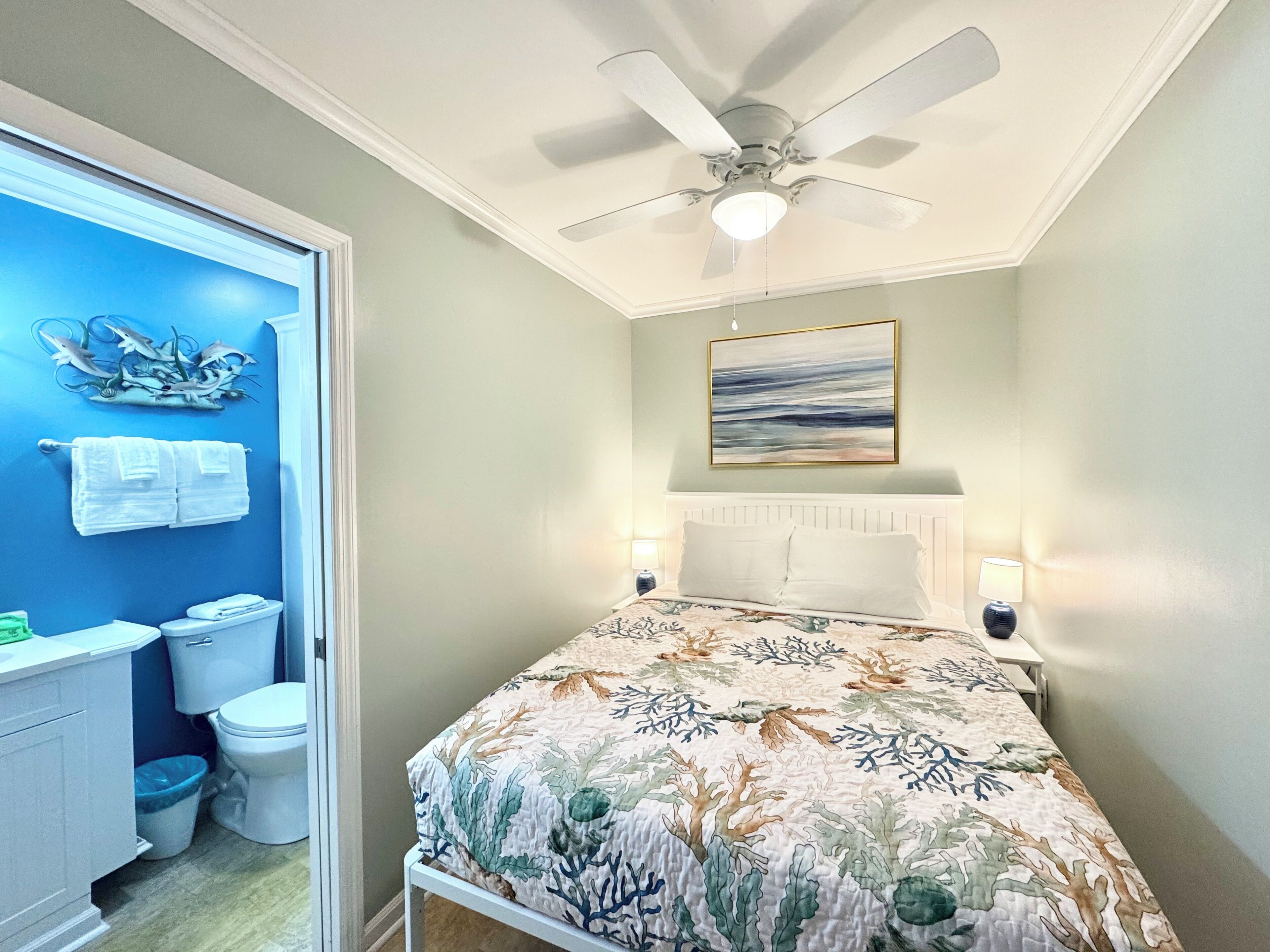 A bedroom with a bed, a ceiling fan, a painting, and a bathroom with a toilet and a trash can.
