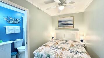 A bedroom with a bed, a ceiling fan, a painting, and a bathroom with a toilet and a trash can.