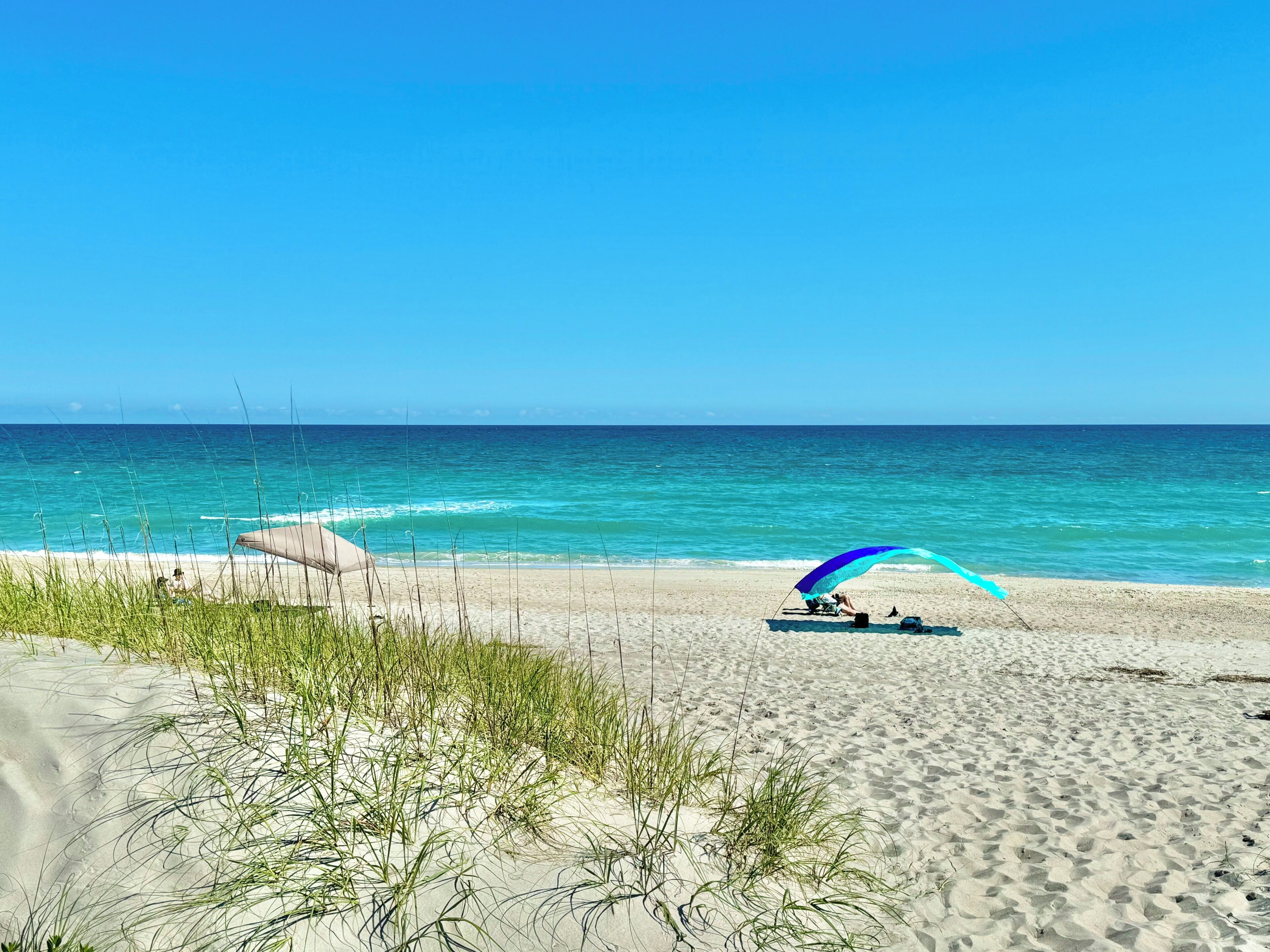 A beach with a clear sky, calm sea, and a person under a blue and white umbrella.