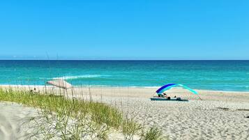 A beach with a clear sky, calm sea, and a person under a blue and white umbrella.