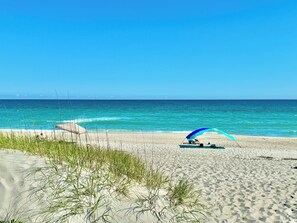 A beach with a clear sky, calm sea, and a person under a blue and white umbrella.