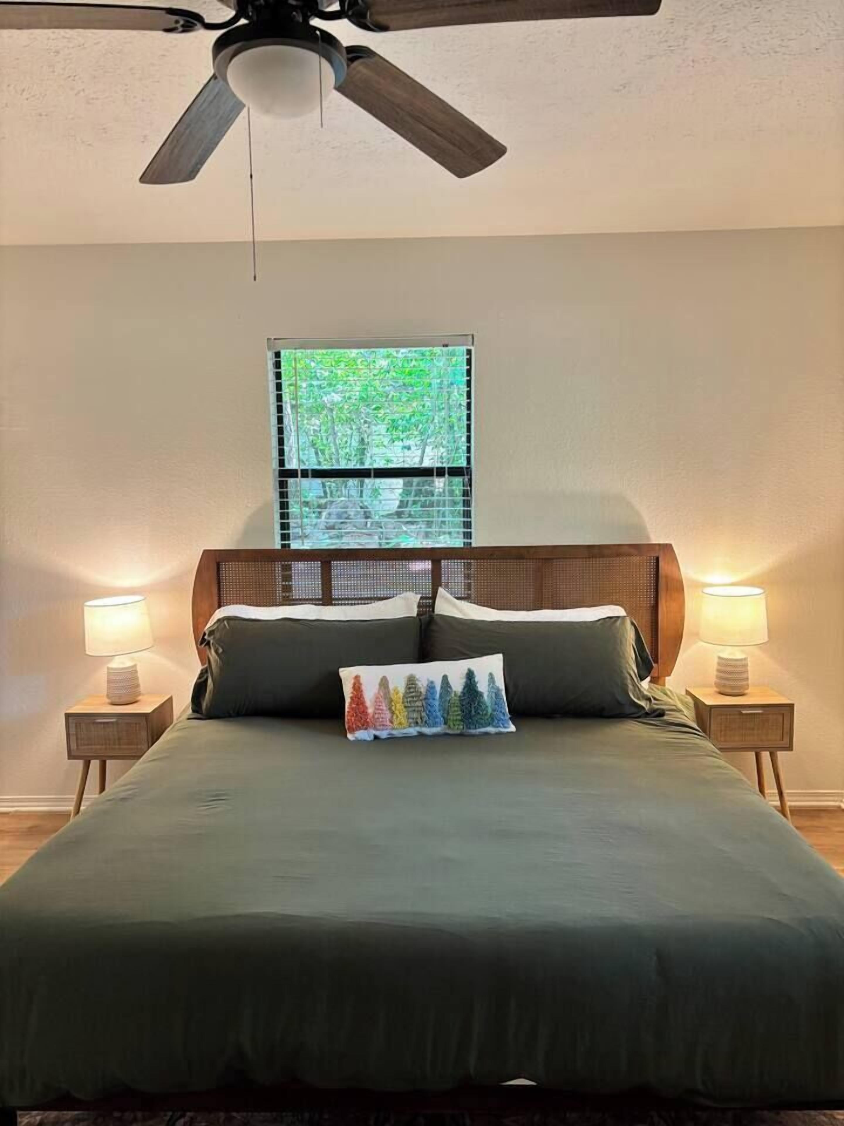 A bedroom with a bed, two bedside lamps, a ceiling fan, and a window with a view of greenery.