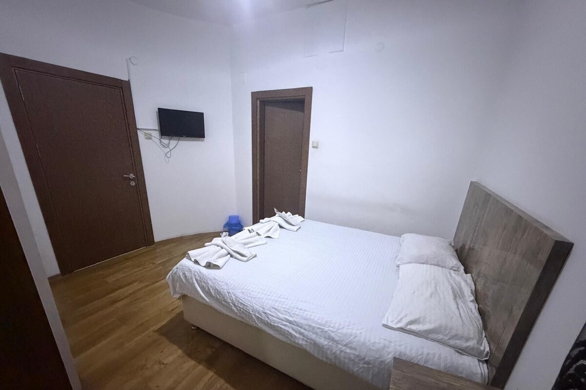 Un dormitorio con cama, cabecera de madera, mesita de noche, televisor montado en la pared y una puerta.