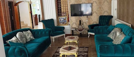 A spacious living room with teal sofas, a chandelier, a flat-screen TV, and a patterned rug.