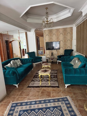 A spacious living room with teal sofas, a chandelier, a flat-screen TV, and a patterned rug. - Kapadokya Pansiyon (Nevşehir)