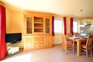 A room with a wooden cabinet with glass doors, a television on a stand, a dining table with chairs, and a kitchen area with cabinets and a window.