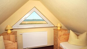 A bedroom with a sloped ceiling, a window with a view of the sea, a radiator, wooden furniture, and two bedside lamps. - Hus Utblick in Fuhlendorf (Fuhlendorf)
