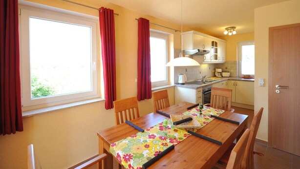 A dining area with a wooden table, chairs, and a floral tablecloth. There is a window with red curtains, a kitchen area with cabinets and a stove, and a door. - Hus Utblick in Fuhlendorf (Fuhlendorf)